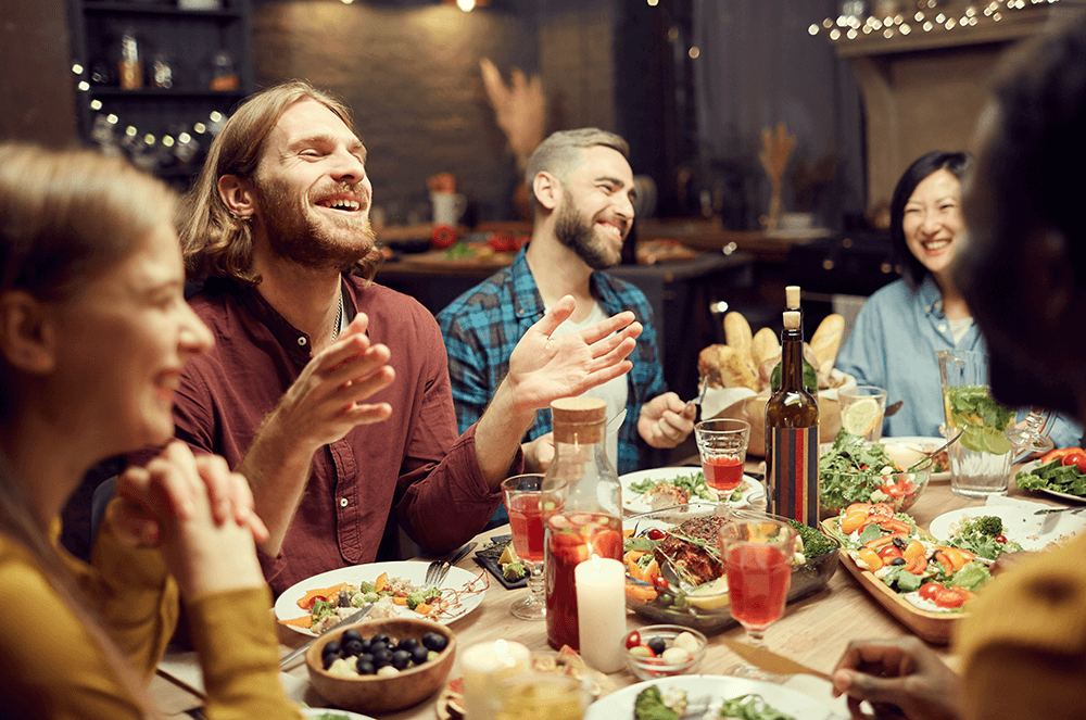 Group of people laughing while gathered at table with food and drinks