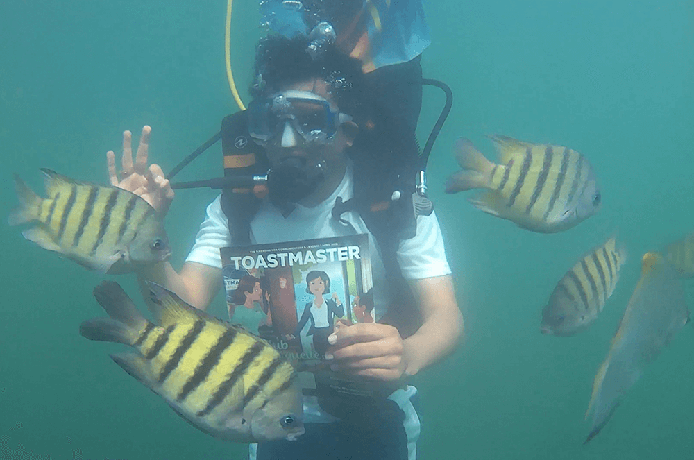 Man scuba diving with Toastmaster magazine signaling he’s safe