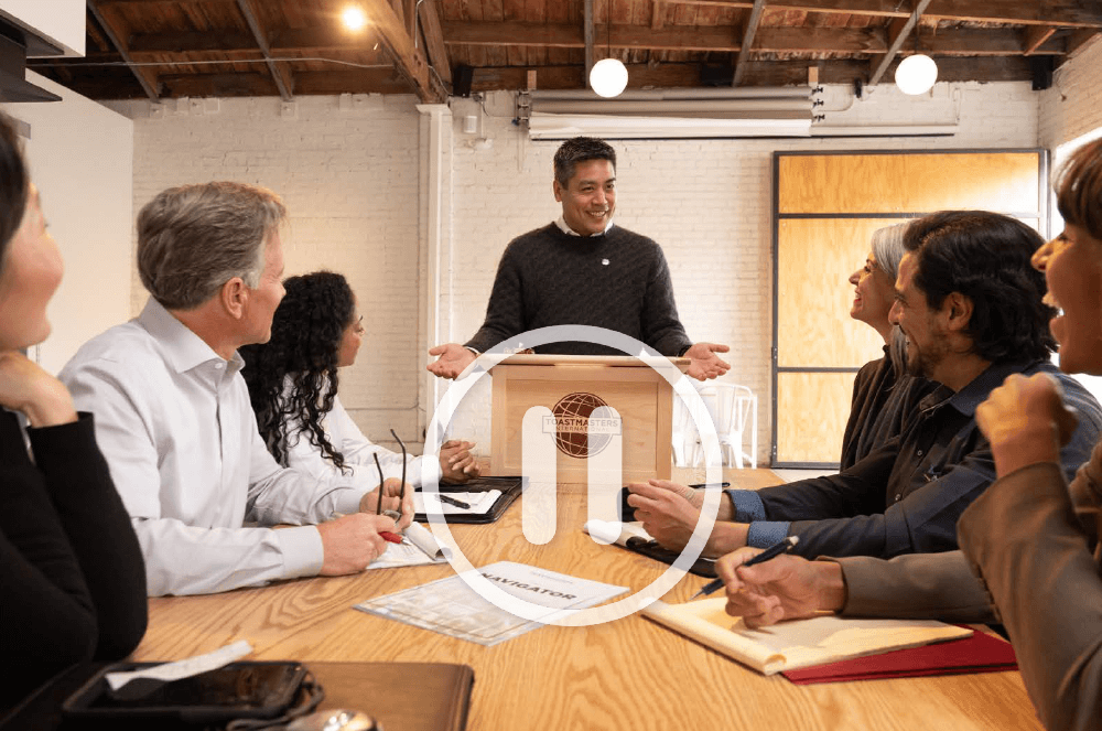 Man speaking at lectern while audience listens around table with white pause icon