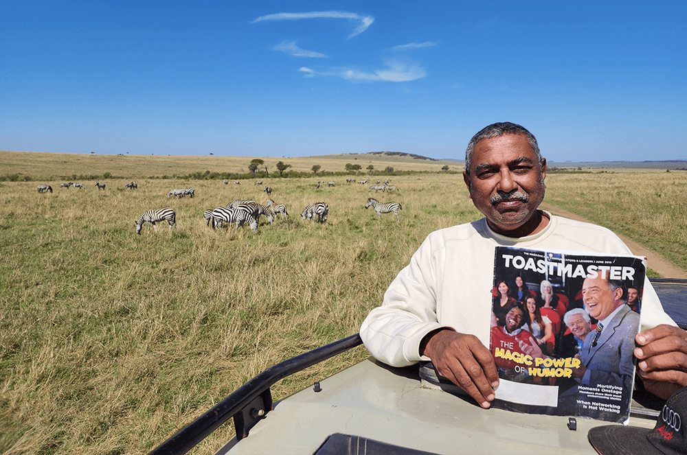 Man holding Toastmaster print magazine with herd of zebras in background