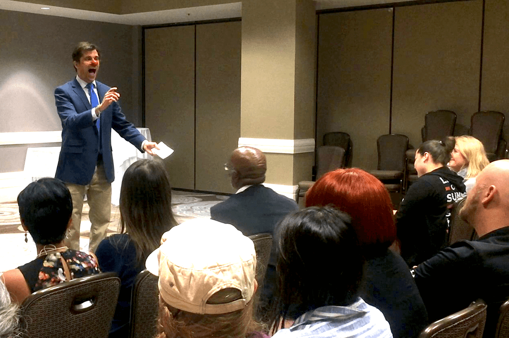 Man speaking to audience wearing blue suit jacket and tie and red clown nose