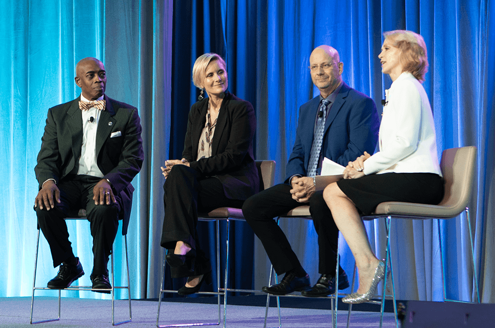 Panel of two men and two women sitting in chairs onstage with blue background