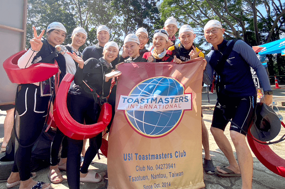 Group of people in wetsuits posing outdoors with Toastmasters banner
