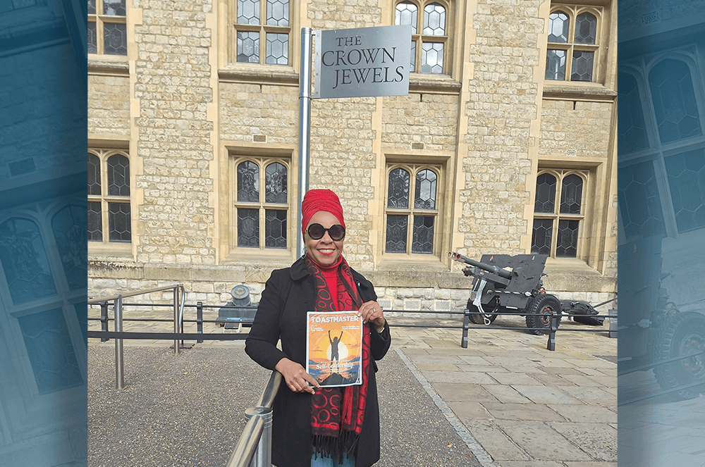 Woman in red head scarf posing with Toastmaster magazine cover in front of the Tower of London