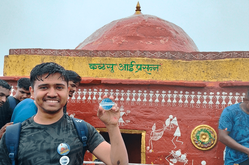 Man holding Toastmasters-branded magnet in front of building on Kalusbai Peak in India