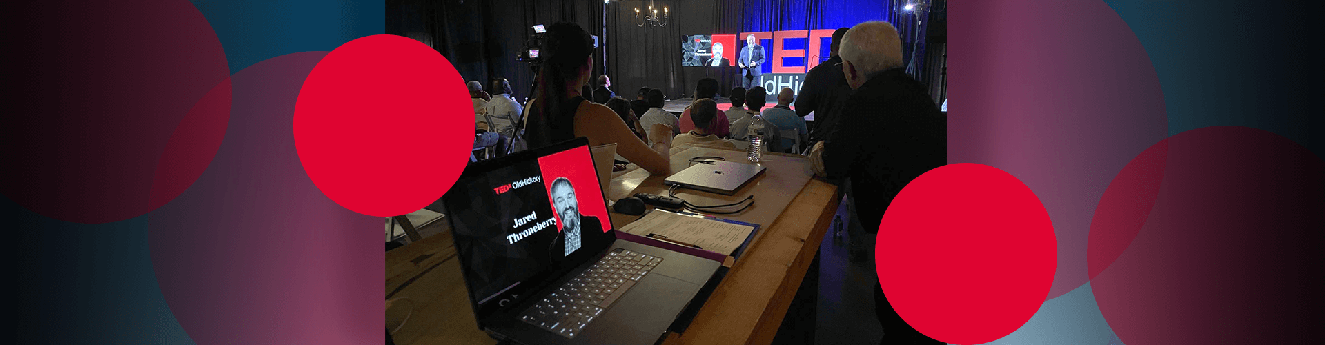 TEDx event with man speaking onstage while audience watches and laptop and notes open on table