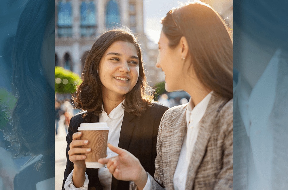 Woman holding coffee cup while speaking to another woman outside with buildings and sun in background