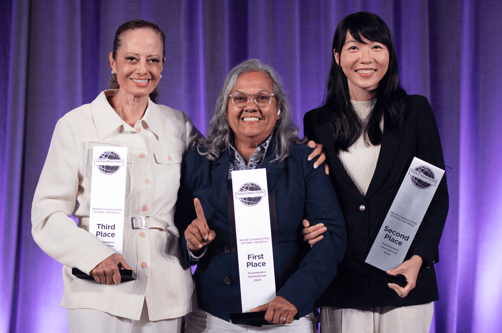 Three women posing onstage holding trophies with purple-lit background