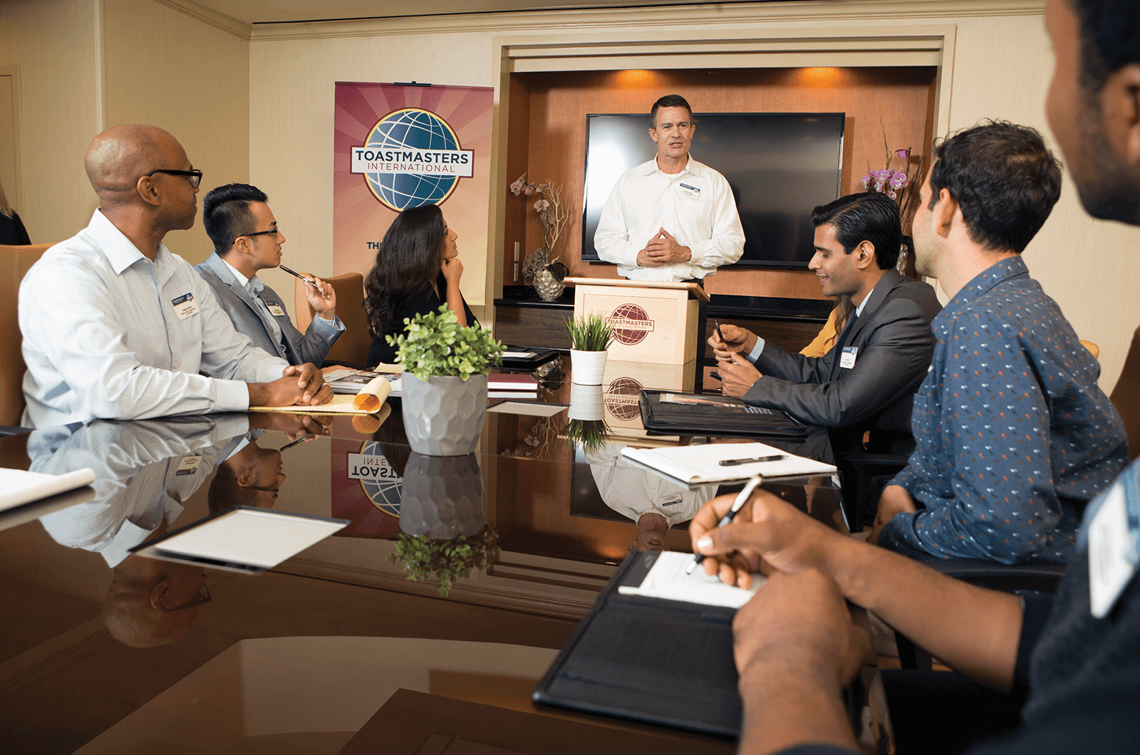 People sitting around table while man speaks at lectern 