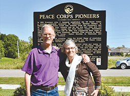 Daniel Grundtner in purple shirt with Ruth Alliband in white scarf in front of Peace Corp sign