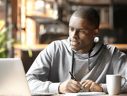 Young man in sweatshirt looking at laptop and taking notes on paper at home