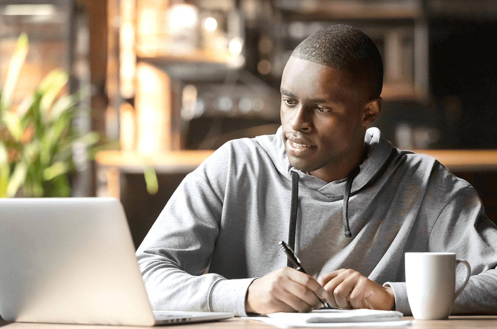 Young man in sweatshirt looking at laptop and taking notes on paper at home
