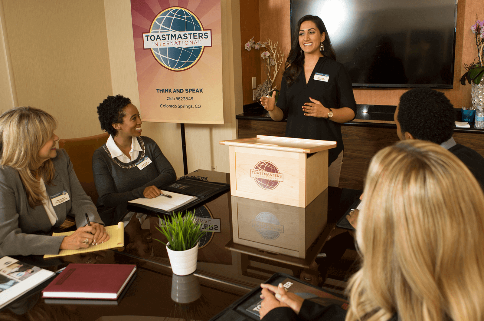 Woman standing behind lectern speaking to people around table