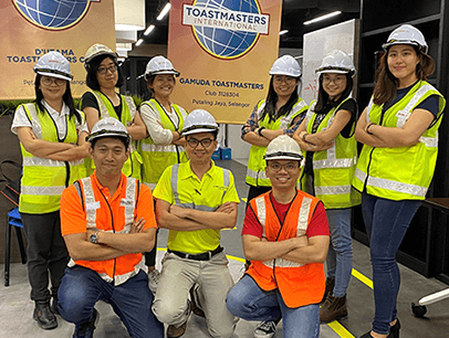 Group of Toastmasters wearing hard hats and yellow and orange vests pose in front of banners