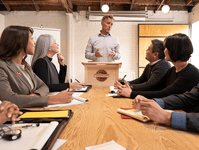 Group of people sitting around conference table while man speaks at a lectern  