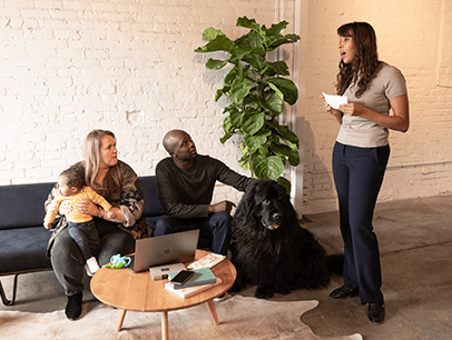Woman holding note card speaking while woman with baby, a man, and dog watch from couch