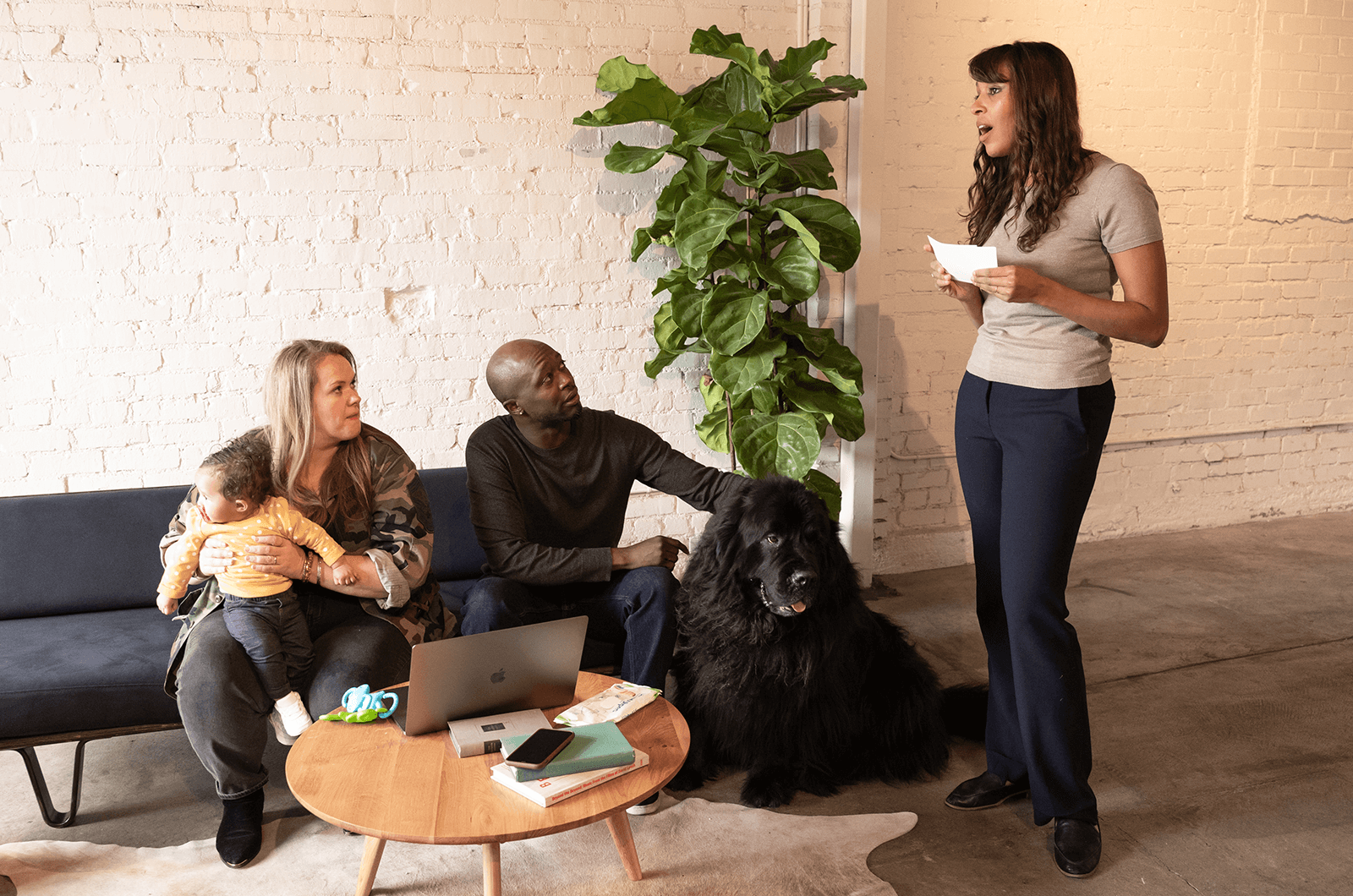 Woman holding note card speaking while woman with baby, a man, and dog watch from couch