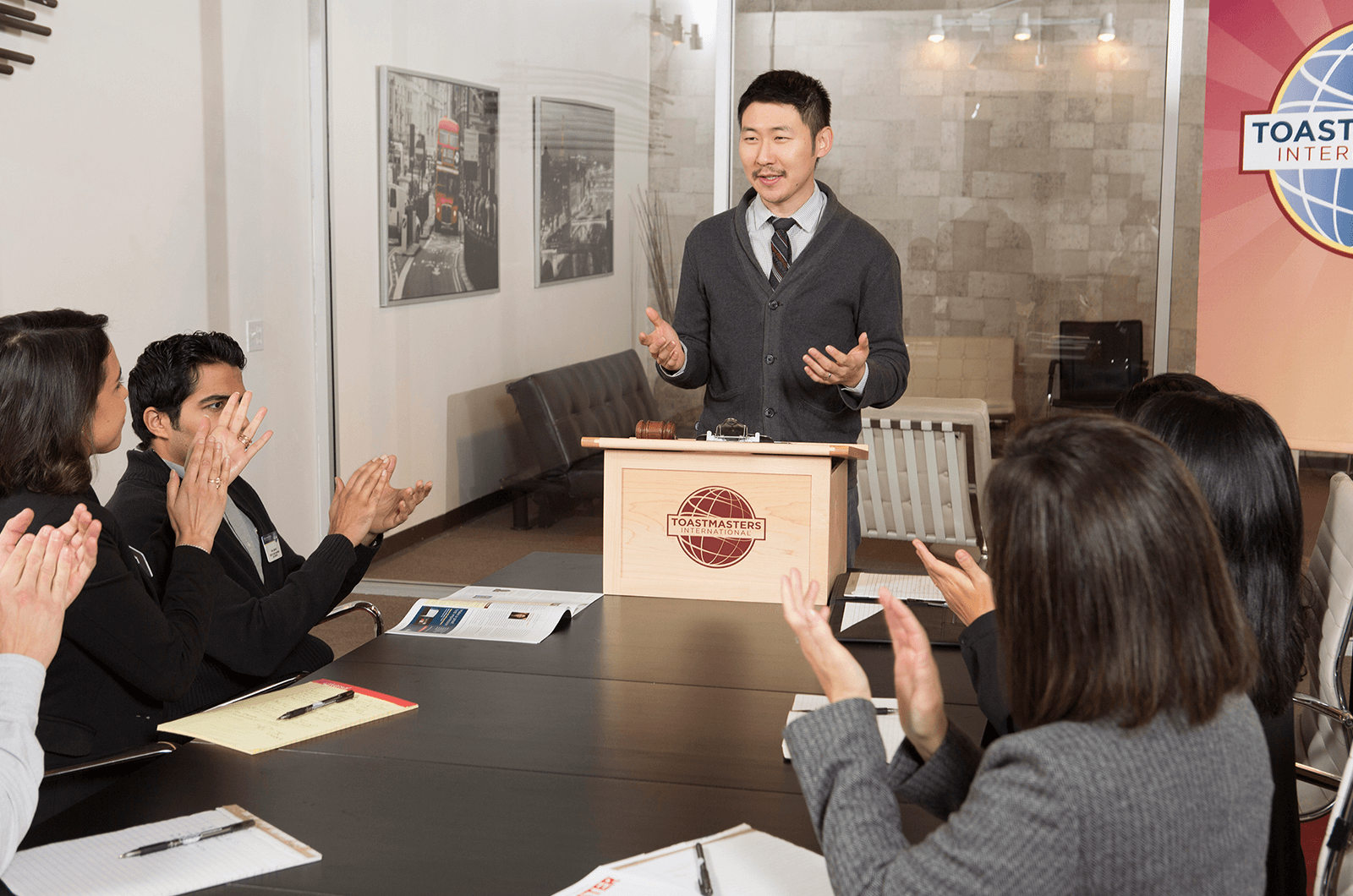 Man standing at lectern speaking to group of people clapping